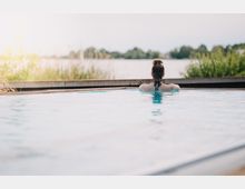 Eine Person sitzt in einem Außenpool und schaut auf eine idyllische Landschaft mit Bäumen und einem See im Hintergrund. Das Wasser des Pools spiegelt den hellen Himmel wider, und am Rand sind Pflanzen sichtbar.