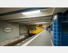 U-Bahn-Station Uhlandstraße in Berlin mit einem gelben Zug der Linie U1, der an einem schmalen Bahnsteig hält. Die Wände sind mit hellen Fliesen verkleidet, und an der rechten Seite befindet sich eine dunkelblaue geflieste Wand.