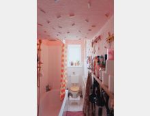 Ein kleines Badezimmer mit einer Badewanne links, die durch einen Duschvorhang mit orangefarbenem Muster abgetrennt ist. Im Hintergrund befindet sich ein Fenster über einer Toilette mit einem Holzklo-deckel; Regale rechts sind mit Pflegeprodukten und Handtüchern bestückt.