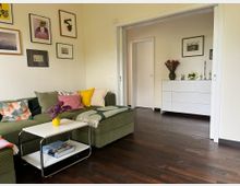 Wohnzimmer mit dunklem Holzboden, grünem Ecksofa mit bunten Kissen und kleinem weißem Beistelltisch. An der Wand hängen mehrere gerahmte Bilder; durch eine breite Schiebetüröffnung ist ein angrenzender Raum mit weißer Kommode und Dekoration zu sehen.