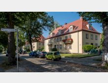 Straßenszene mit zwei- bis dreistöckigen Wohngebäuden im traditionellen Stil, cremefarbene Fassaden mit roten Ziegeldächern und Blumenkästen an den Fenstern. Kopfsteinpflasterstraße mit parkenden Autos und Bäumen im Vordergrund, Straßenschild „Margaretenstraße“ links im Bild.