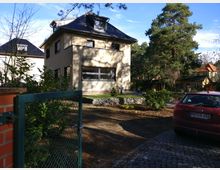 Einfamilienhaus mit heller Fassade und dunkel gedecktem Dach in einer grünen, bewaldeten Umgebung. Im Vordergrund ein offenes grünes Metalltor, ein rotes Auto auf einem gepflasterten Weg und gepflegte Gartenanlagen.