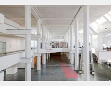 Großer, heller Innenraum eines modernen Gebäudes mit weiß gestrichenen Säulen, hohen Decken und großen Fensterfronten, die viel Tageslicht hereinlassen. In der Mitte befindet sich ein Schild mit der Aufschrift „Garderobe / Cloakroom“, und eine Treppe führt zu einer erhöhten Ebene.