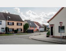 Straßenszene in einem Wohngebiet mit Einfamilienhäusern, teilweise verputzt und mit Satteldächern. Rechts im Vordergrund ein kleines Geschäft mit der Aufschrift "Friseursalon Brüggemann", daneben ein Gehweg und Verkehrsschilder.