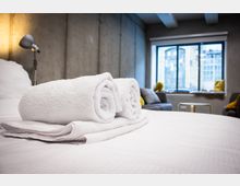 Modernes Hotelzimmer im Industrial-Stil mit Betonwänden, großem Fenster zur Stadt und einem Bett im Vordergrund, auf dem zwei gerollte weiße Handtücher liegen. Im Hintergrund sind ein Sofa mit Kissen, eine Stehlampe und ein kleiner Tisch zu sehen.