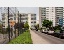 Wohngebiet mit mehreren Hochhäusern aus Beton mit Balkonen; im Vordergrund ein Gehweg, eine schmale Straße mit parkenden Autos, grüne Rasenflächen und ein umzäunter Bereich auf der linken Seite. Bäume und Büsche umgeben die Gebäude.