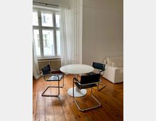 Ein heller Raum mit Holzdielenboden, einem runden weißen Tisch und drei Stühlen mit Metallrahmen und schwarzem Leder. Im Hintergrund befindet sich ein großes Fenster mit weißen Vorhängen und ein Heizkörper, rechts steht ein heller Sofaabschnitt.