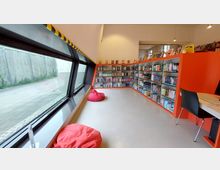 Moderner Leseraum mit großen, geneigten Fenstern auf der linken Seite, durch die grünes Außengelände sichtbar ist. Auf der rechten Seite befinden sich orangefarbene Bücherregale mit einer Vielzahl von Büchern, zwei rote Sitzsäcke stehen auf dem hellen Boden, und ein Tisch mit einem Stuhl ist im Vordergrund zu sehen.