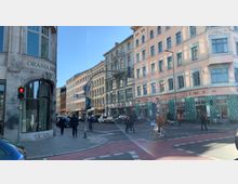 Straßenszene in einer Stadt mit historischen Wohnhäusern und Geschäften. Im Vordergrund eine rote Fußgängerampel, ein Fahrradfahrer sowie Passanten auf einem Gehweg; im Hintergrund Geschäfte wie "Café Alibi" und "Dante Connection".