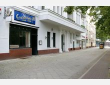 Straßenszene in einer Wohngegend mit einem weißen Gebäude, das eine Cocktailbar mit blauem Schild sowie Eingangsbereiche mit Säulen umfasst. Der Gehweg besteht aus Kopfsteinpflaster und ist von Bäumen und weiteren Gebäuden gesäumt.