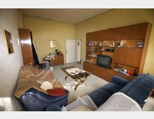 Wohnzimmer mit gelber Wandfarbe, ausgestattet mit einer großen Holz-Schrankwand, einem Röhrenfernseher, zwei Sofas und einem Couchtisch mit Glasplatte. Im Raum befinden sich persönliche Gegenstände und Dekorationen, die eine bewohnte, ältere Einrichtung widerspiegeln.