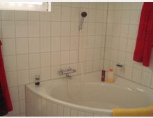 Weiß gefliestes Badezimmer mit einer Eckbadewanne, einer Handbrause und Ablageflächen mit Shampoo- und Duschflaschen. Ein rotes Handtuch hängt links an der Wand, ein gelbes Handtuch liegt über dem Wannenrand.