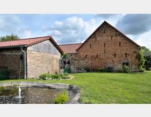 Ein rustikaler Hof mit zwei Gebäuden aus Ziegelstein und roten Dachziegeln, umgeben von Rasenfläche. Im Vordergrund befindet sich ein kleiner Wasserteich mit einer Fontäne.