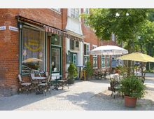 Straßenszene mit einem Café vor einem roten Backsteingebäude, ausgestattet mit weißen Fenstern und grün-weißen Türen. Im Außenbereich stehen Tische und Stühle auf einem gepflasterten Gehweg, teilweise beschattet durch Sonnenschirme und kleine Topfpflanzen.