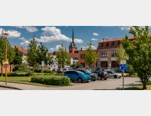 Straßenszene mit einem kleinen Parkplatz, umgeben von Bäumen und Gebäuden mit roten Ziegeldächern. Im Hintergrund ist eine Kirche mit einem spitzen Turm zu sehen, während Verkehrsschilder und parkende Autos den Vordergrund dominieren.