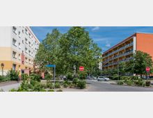 Wohngebiet mit mehrstöckigen Wohnhäusern, links ein Gebäude in Weiß und Gelb, rechts eines mit oranger Fassade und Balkonen. In der Mitte der Straße stehen Bäume und Verkehrsschilder, umgeben von parkenden Autos und Grünanlagen.