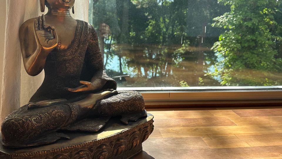 Bronzefarbene Buddha-Statue auf einem Holzboden direkt vor einer großen Glasfensterfront mit Vorhang. Durch das Fenster sind ein sonniger Garten mit Bäumen und ein Gewässer im Hintergrund zu sehen.