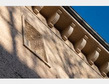 Fassade eines Gebäudes mit grober, sandfarbener Struktur und schattenspendenden Verzierungselementen unterhalb des Daches, sowie einem kleinen, gefalteten Lüftungsgitter in einer Vertiefung. Der blaue Himmel ist im Hintergrund sichtbar.