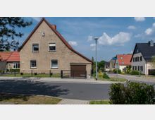 Ein Wohngebiet mit Einzelhäusern, darunter ein hellbraunes Haus mit Satteldach und angegliederter Garage. Die Straße mit Gehwegen und einer Laterne führt durch die Szene, während im Hintergrund weitere Häuser mit gepflegten Gärten sichtbar sind.