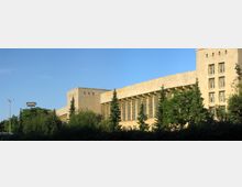 Großes, beige-steinernes Gebäude mit klaren Linien und hohen Fenstern, flankiert von Bäumen und umgeben von einer grünen Vegetation. Der Himmel ist wolkenlos und hellblau.