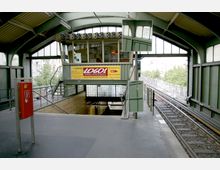 Ein erhöht gelegener S-Bahnhof mit einer überdachten Plattform aus grünem Stahl und Glas. Links befindet sich ein roter Notrufkasten, in der Mitte ein Treppenabgang, und rechts die Gleise mit einer einfahrenden Bahnstrecke.