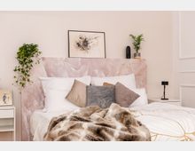 Ein modernes Schlafzimmer mit einem Bett, dessen Kopfteil aus rosa Marmor-Optik besteht. Das Bett ist mit weißen Kissen, grauen und beigen Dekokissen sowie einer gemusterten Decke dekoriert, umgeben von weißen Nachttischen, Pflanzen und einer abstrakten Wandkunst.