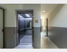 Breiter Flur in einem modernen Gebäude mit hellen Wänden und einem offenen Aufzug mit Edelstahlverkleidung. Rechts führt der Korridor zu weiteren Türen, und an der Wand ist ein Notfallschalter zu sehen.