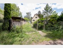 Eingezäuntes Grundstück mit Drahtzaun und Tor, dahinter ein schmaler, grasbewachsener Weg zu einem kleinen, hellen Einfamilienhaus mit steilem Giebeldach. Das Gelände ist dicht bewachsen mit Bäumen, Sträuchern und einem überdachten Holzunterstand links.