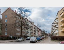 Straßenszene mit mehrstöckigen Wohnhäusern auf beiden Seiten, teils mit Balkonen, und einer Reihe von geparkten Autos am Straßenrand. Die Bäume ohne Blätter und der wolkige Himmel deuten auf eine Winter- oder Frühjahrsstimmung hin.