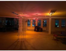 Ein leerer Raum mit großen Fenstern, die nächtliche Stadtlichter zeigen. Im Raum befinden sich eine beleuchtete, transparente Säule in der Mitte, eine dunkle, skulpturartige Sitzgelegenheit, einzelne Hängedekorationen und ein Tisch mit einem roten Stuhl.