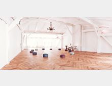 Heller Raum mit Holzboden in Fischgrätenmuster, weiß gestrichenen Wänden und freiliegenden Balken an der Decke. Auf dem Boden sind runde Sitzkissen verteilt, und ein Kronleuchter hängt zentral von der Decke.