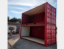 Zwei rote Frachtcontainer sind übereinander gestapelt, wobei der untere Container an der Vorderseite offen ist. Im Hintergrund sind ein niedriges Gebäude mit der Aufschrift "Antik" sowie einige Bäume und dekorative Elemente zu sehen.