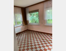 Ein leerer Raum mit rosa Wänden, zwei großen Fenstern mit weißen Gardinen und gelben Vorhängen. Der Boden ist mit rot-weißen Schachbrettfliesen bedeckt, und ein Heizkörper befindet sich unter einem der Fenster.