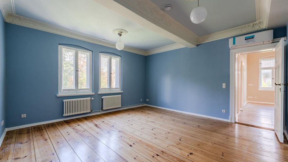 Leerer Wohnraum mit blau gestrichenen Wänden, breiten Holzdielen und zwei hohen Fenstern über weißen Heizkörpern. Stuckverzierte Decke mit Kugelleuchten und eine Türöffnung führt in einen hellen Nebenraum.