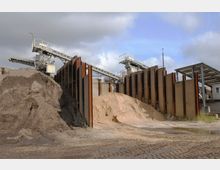 Industriegelände mit großen Sandhaufen und einer Förderanlage aus Stahl. Im Hintergrund befinden sich vertikale Stahlträger sowie eine Überdachung mit einer metallischen Konstruktion.