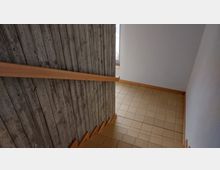 Treppenhaus mit hellbraunen Fliesen, einer Sichtbetonwand auf der linken Seite und einem hölzernen Handlauf. Rechts sind weiß gestrichene Wände sichtbar, mit einem Fenster im Hintergrund.