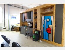 Eine moderne Büroküche mit einer hölzernen Einbauwand, die einen farbig bemalten Kühlschrank, Regale mit Gläsern und eine Kaffeemaschine enthält. Im Hintergrund sind weiße Fliesenwände, ein Schreibtisch mit Stuhl und helle Fenster zu sehen.