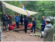 Eine Gruppe von Menschen steht in einem Wald unter einem großen, beigefarbenen Sonnensegel, das mit bunten Stoffflaggen dekoriert ist. Auf dem Boden befinden sich Teppiche und Kissen, umgeben von Bäumen und Natur.