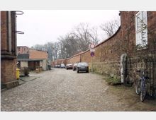 Eine gepflasterte Straße führt entlang einer hohen, historischen Backsteinmauer. Auf der linken Seite sind einige Backsteingebäude und geparkte Autos zu sehen, während ein Fahrrad neben der Mauer auf der rechten Seite steht. Im Hintergrund sind Bäume und weitere Mauern sichtbar.