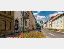 Straßenszene in einer Wohngegend mit Reihen von historischen und modernisierten Mehrfamilienhäusern, teils mit Balkonen. Am Straßenrand sind rote Mohnblumen und andere Wildblumen zu sehen, während ein Auto geparkt ist; der Himmel ist klar mit ein paar Wolken.