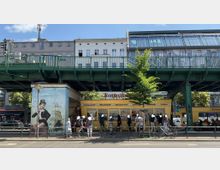 Straßenansicht mit einem gelben Imbissstand unter einer grünen Hochbahntrasse. Im Vordergrund stehen Menschen in einer Warteschlange, daneben ein gemaltes Wandbild und ein Baum. Dahinter sind Wohn- und Bürogebäude sichtbar.