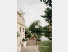 Holzterrasse an einem Flussufer neben einem hellen Gebäude mit Geländer, Liegestühlen und großen Pflanzkübeln. Daneben führt eine Rasenfläche zum ruhigen Wasser, umgeben von Bäumen, mit einem Schild „Land’s End“ am Ufer.