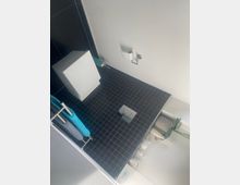 Blick von oben in ein kleines, gefliestes Badezimmer mit dunklen Bodenfliesen, bodengleicher Dusche mit Ablaufrinne und Glasabtrennung. An der Wand sind eine Toilettenpapierhalterung und eine kompakte weiße Sanitärinstallation zu sehen, daneben ein Handtuchhalter mit blauem Handtuch.