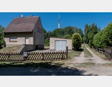 Einfamilienhaus mit hellem Putz und Satteldach, daneben eine freistehende Garage mit Metalltor. Im Vordergrund ein Holzzaun und ein gepflasterter Weg, dahinter Bäume und ein Mobilfunkmast.