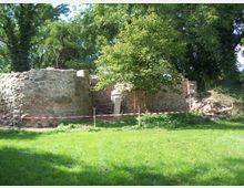 Ruine einer alten Steinmauer inmitten einer grünen Wiese, umgeben von Bäumen. Vor der Ruine ist der Bereich mit einem rot-weißen Absperrband markiert.