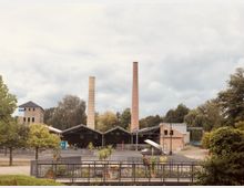 Industriekomplex mit historischen Backsteingebäuden und zwei hohen Schornsteinen, umgeben von Bäumen und freier Fläche; im Vordergrund ein Geländer und vereinzelte Pflanzen.