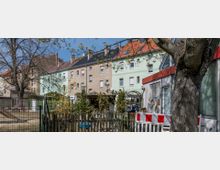 Wohngebiet mit mehrstöckigen Reihenhäusern in hellen Pastellfarben, umgeben von einem kleinen Garten mit Bäumen, Sträuchern und einem Holzzaun. Im Vordergrund steht ein großer Baum, und ein Spielplatz mit einer Rutsche ist teilweise sichtbar.