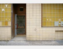 Eine Eingangstür in eine Gebäudefassade aus beigefarbenen Kacheln mit gelben, vergitterten Fenstern. Die Tür hat ein dekoratives, gitterartiges Metallmuster, einen roten Griff und eine Lüftungsöffnung darüber; der Gehweg davor ist uneben.