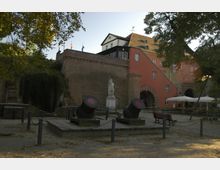 Ein historischer Hof mit Ziegelmauern, einer Statue auf einem Sockel in der Mitte und zwei großen Kanonen im Vordergrund. Im Hintergrund ein Gebäude mit roter Fassade, mehreren Fenstern und Rundbögen sowie ein Baum, der teilweise den Blick beschattet.
