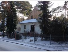 Einfamilienhaus in einer verschneiten Umgebung, mit weißer Fassade, Spitzdach und Holzelementen im oberen Bereich. Vor dem Haus befinden sich ein Metallzaun, hohe Bäume und ein mit Schnee bedeckter Gehweg.